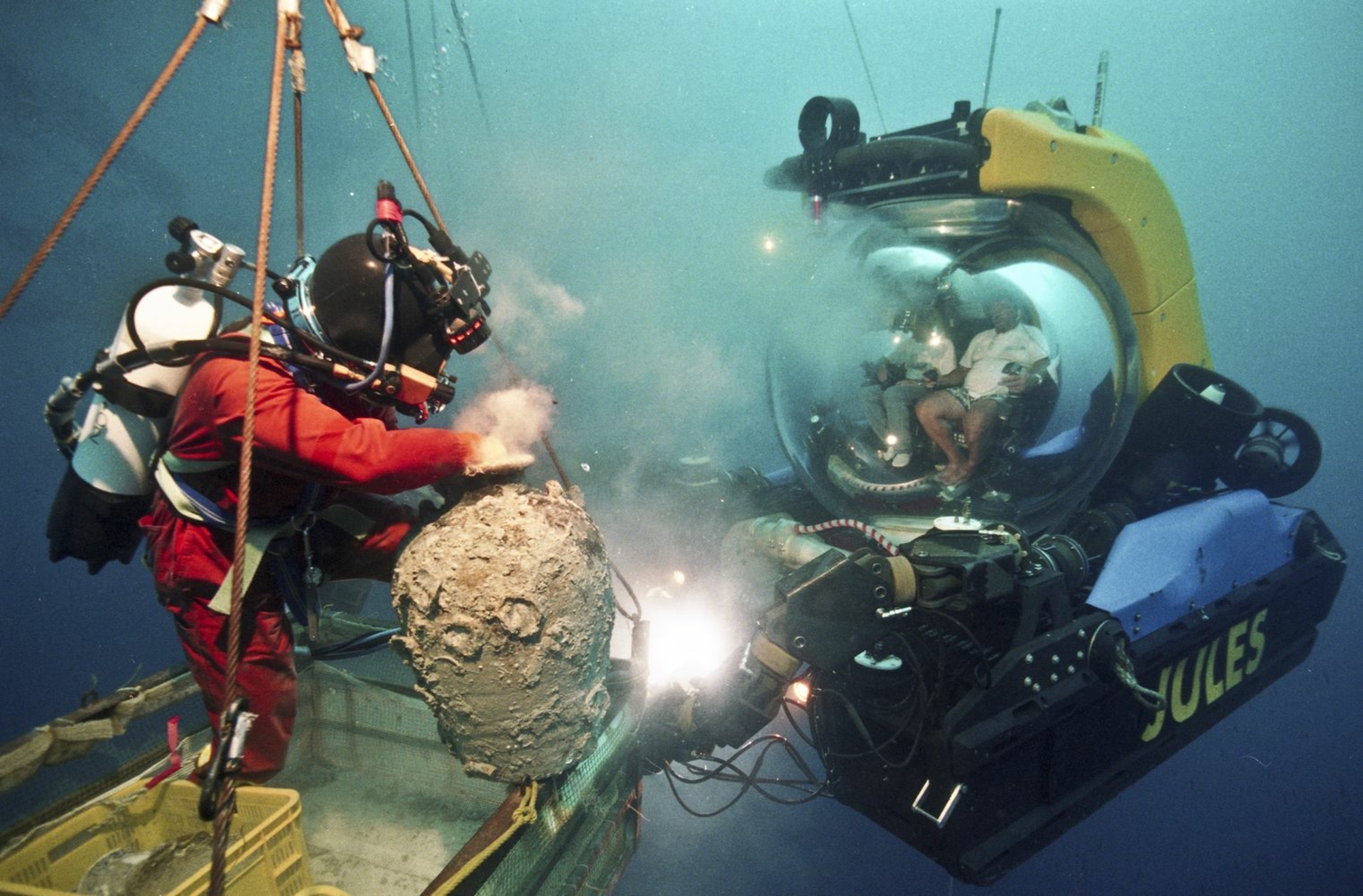 Photographie d'un plongeur tenant un objet observé par deux personnes dans un module sous-marin