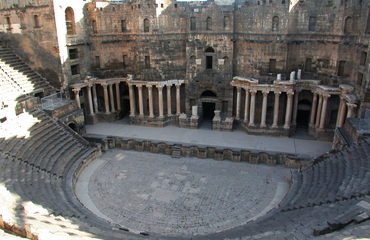 Bosra théâtreVue sur la cavea et le scaenae frons. Ce théâtre est entièrement construit sur un terrain plat. Les gradins, d’une contenance de 8000 à 9000 personnes, sont sur un plan semi-circulaire légèrement outrepassé (102 m de diamètre), ouvrant au nord sur un profond et imposant mur de scène de même hauteur.